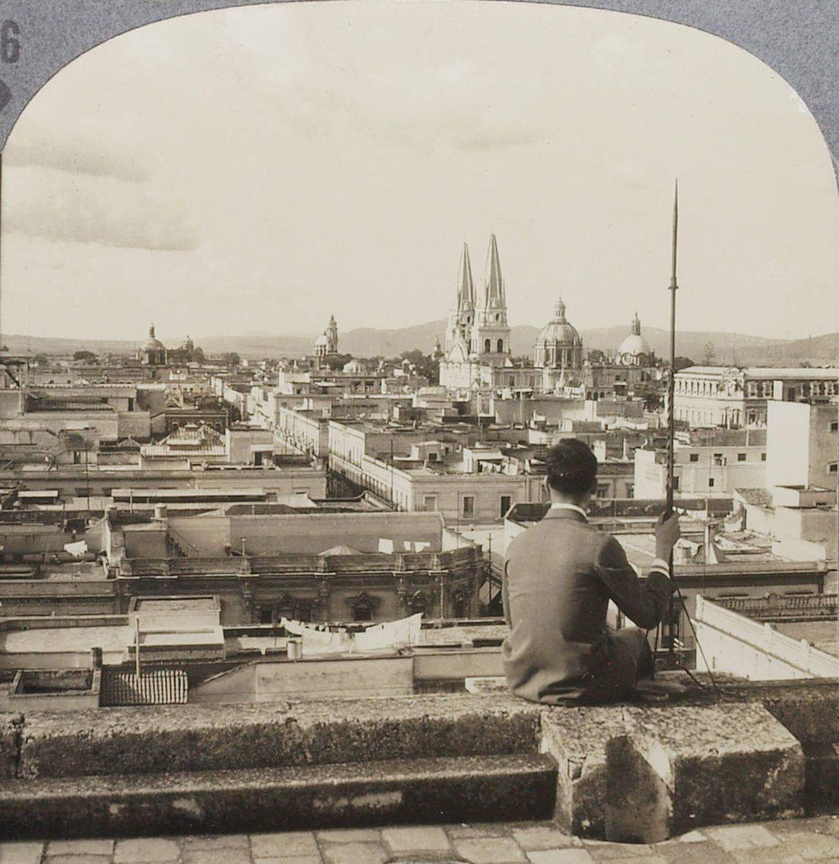 Este es un retrato en blanco y negro de la ciudad de Guadalajara con estilo antiguo, que muestra a un hombre sentado de espaldas en el borde de lo que parece ser una azotea o una terraza alta. El hombre lleva puesto un traje formal con chaqueta, pantalones y zapatos, junto con un sombrero elegante. Parece estar sosteniendo un bastón o una caña con su mano derecha y está mirando hacia el horizonte. La escena captura un paisaje urbano con varios edificios que se extienden hasta el fondo. Es una vista panorámica en la que se destacan dos prominentes catedrales o iglesias con torres puntiagudas y detalles arquitectónicos intrincados. También hay múltiples cúpulas y tejados que indican una densa área urbana. El cielo sobre la ciudad es claro, con algunas nubes dispersas, lo que sugiere que podría ser un día tranquilo. El formato de la imagen es de un rectángulo con los bordes superiores redondeados, lo que le da la apariencia de una vieja fotografía estereoscópica o de tarjeta de visita, que era un estilo común de presentación de fotografías en el siglo XIX y principios del XX. La imagen provoca una sensación de nostalgia y contemplación, como si capturara un momento en la historia de una ciudad desconocida.