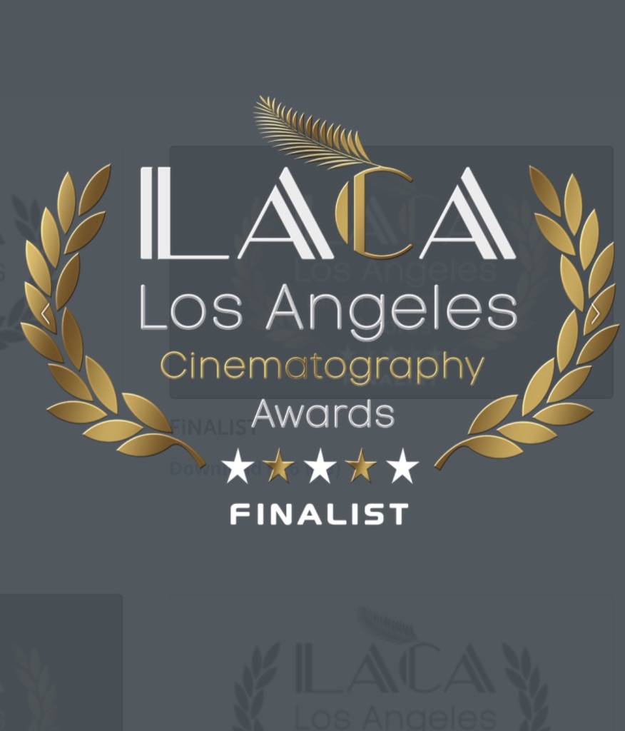 La imagen muestra el logotipo de los Premios de Cinematografía de Los Ángeles (Los Angeles Cinematography Awards, LACA). El logotipo tiene texto en blanco con algunas letras en dorado, rodeado por dos ramas de laurel doradas. Hay una pluma dorada encima en una C. El texto incluye las palabras LACA, Los Angeles, Cinematography Awards y FINALIST, junto con una fila de cinco estrellas con la palabra FINALIST debajo.